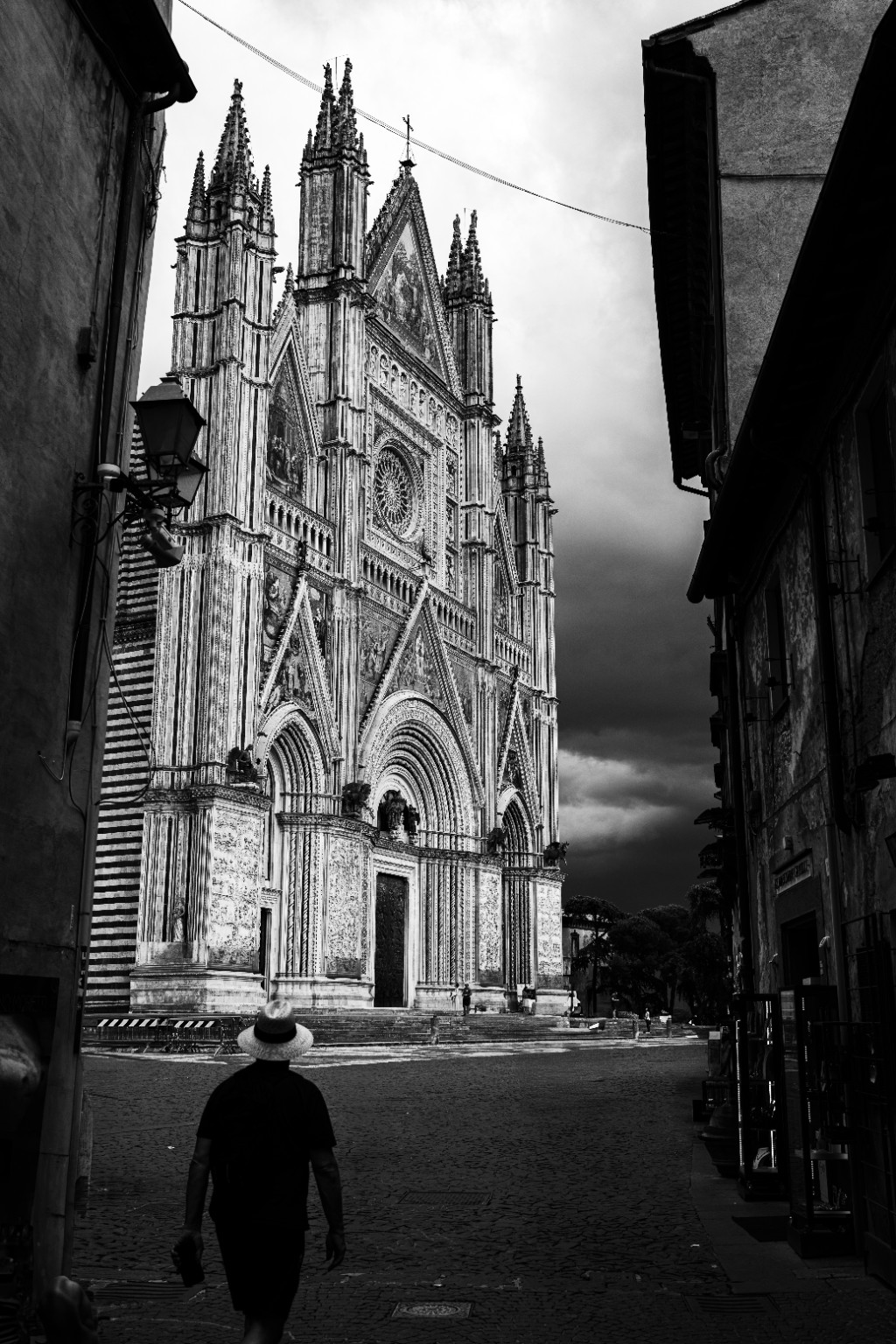 A narrow street opens onto the ornate facade of Orvieto Cathedral, with a passing figure giving scale to its Gothic detail. Orvieto, Umbria, Italy.