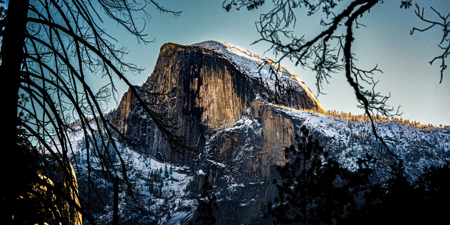 Snow traces Half Dome and the surrounding granite, framed by dark branches and a narrow band of warm light. Yosemite National Park, California.