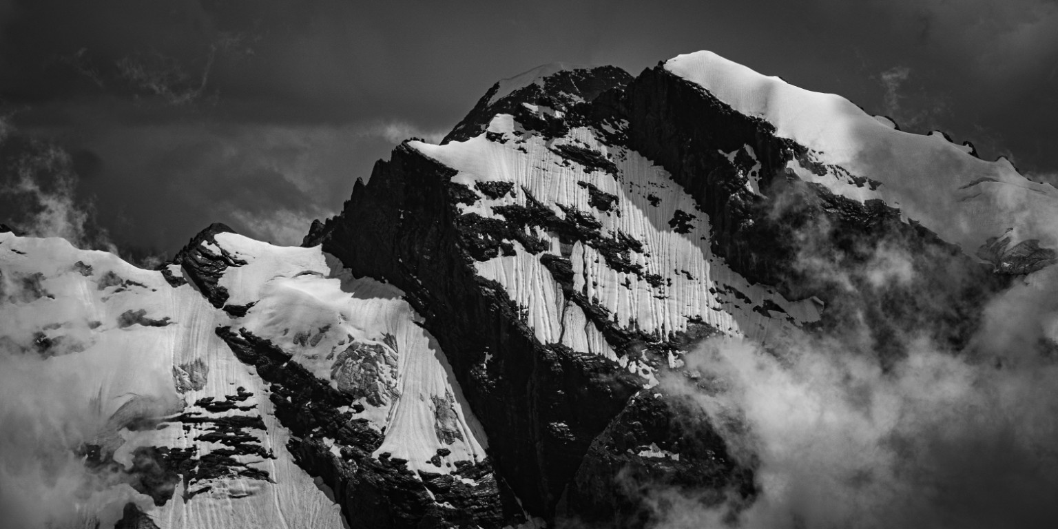 Clouds drift around a snow-covered summit, separating bright ridges from deep rock shadows. Switzerland.