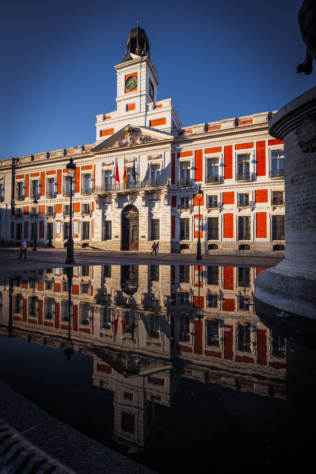 The red-and-white facade of the Real Casa de Correos is reflected in water across Madrid’s Puerta del Sol. Madrid, Spain.