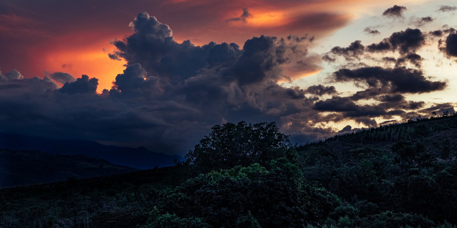 Warm sunset light glows behind sculpted clouds above a dark, wooded mountainside. Near Ceilán, Colombia.
