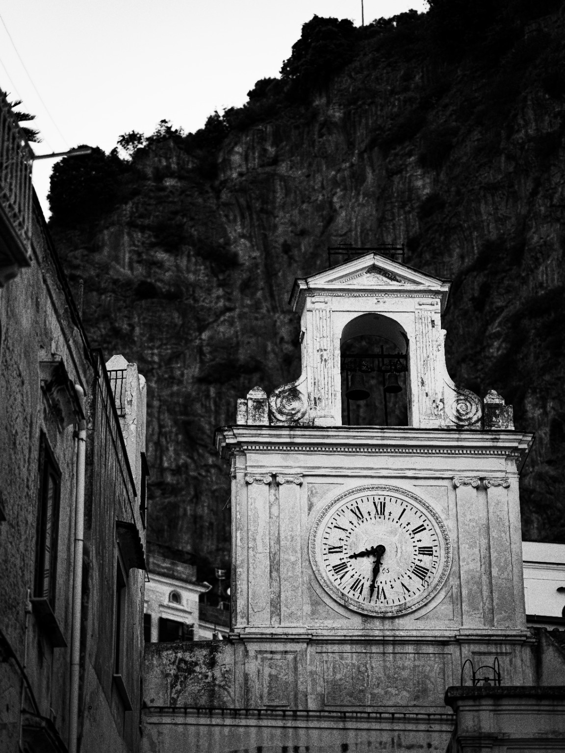 The church clock and open bell arch stand against a steep rock wall, bringing the scale of the cliff into a close architectural view. Atrani, Amalfi Coast, Italy.