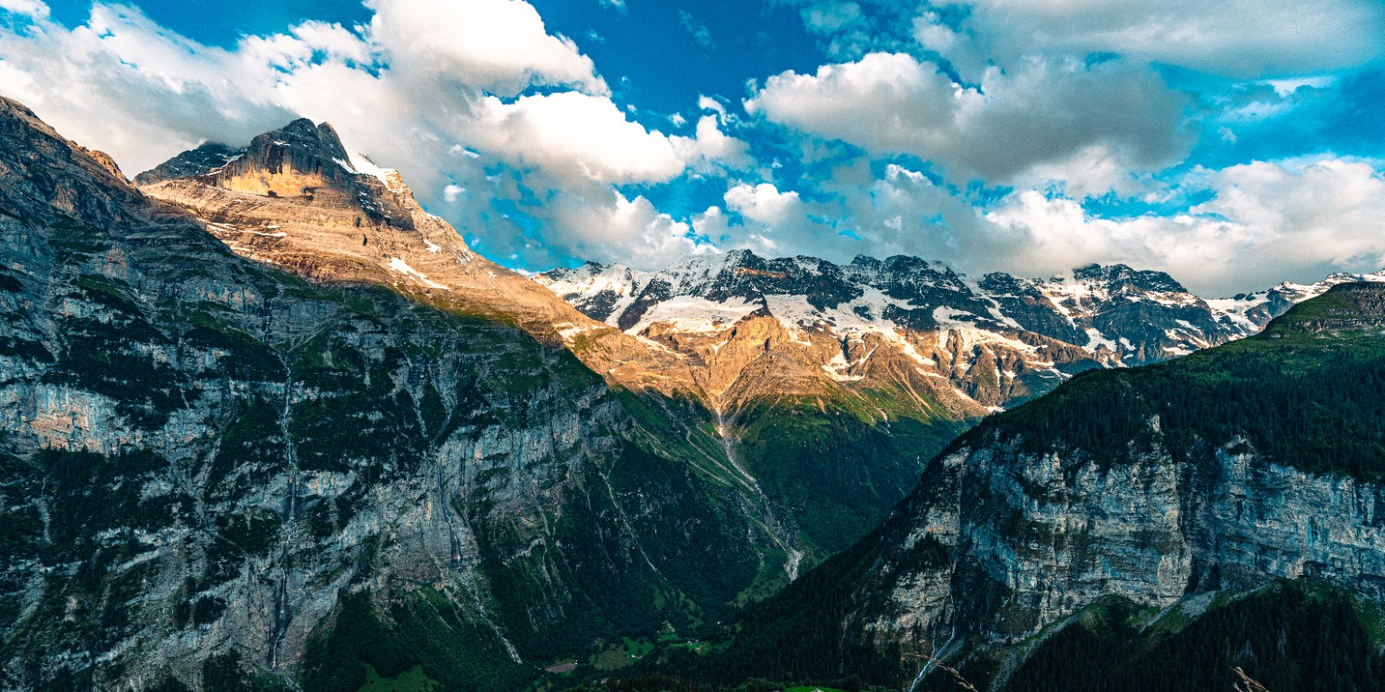 Sunlight crosses the high slopes above a deep valley, with snow and cloud defining the distant ridgeline. Switzerland.