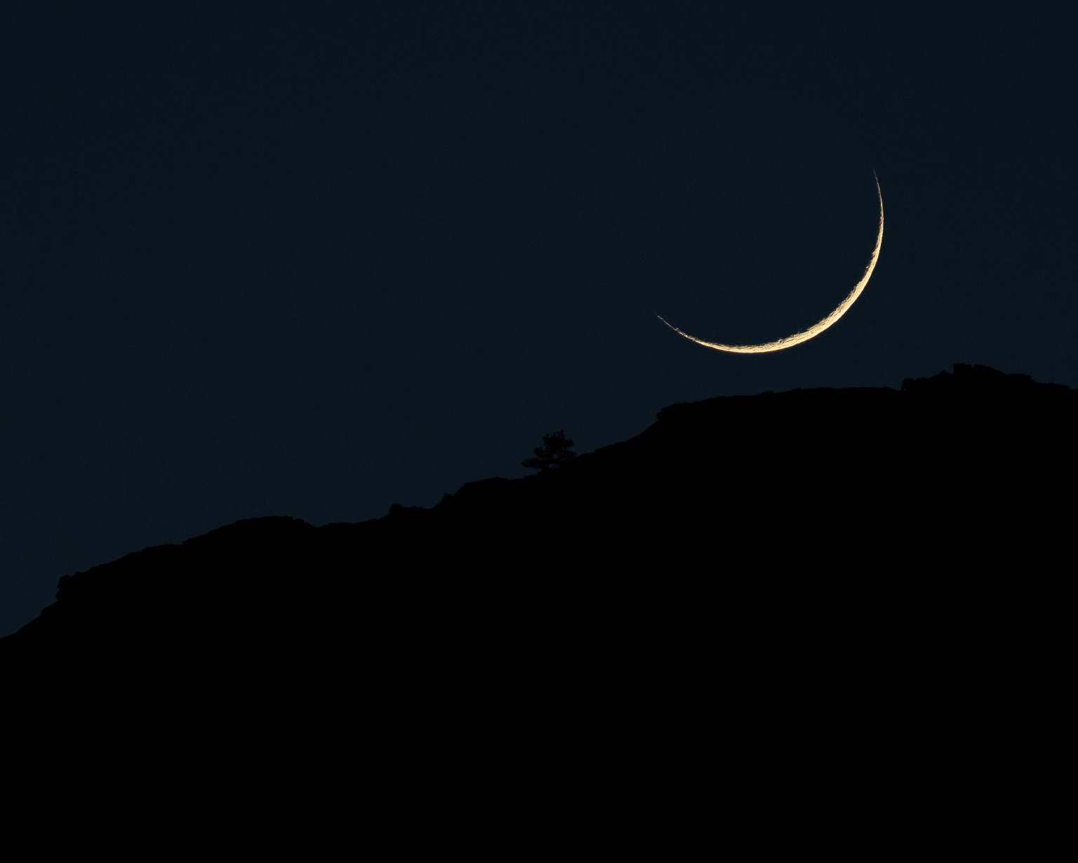 A fine golden crescent hangs above a dark ridgeline, with the landscape reduced to a simple silhouette. Zion National Park, Utah.